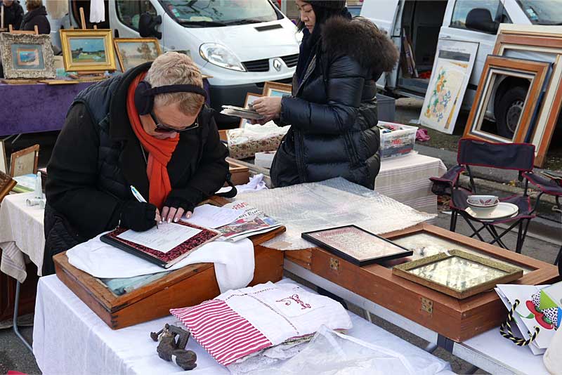 プロヴァンスの蚤の市で領収書を書くフランス人の売り手女性。アンティークの額や布ものなどが店に並んでいる。